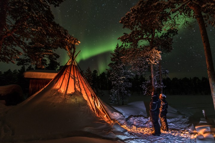 Two people stand near a lit tent under the northern lights in a snowy forest.