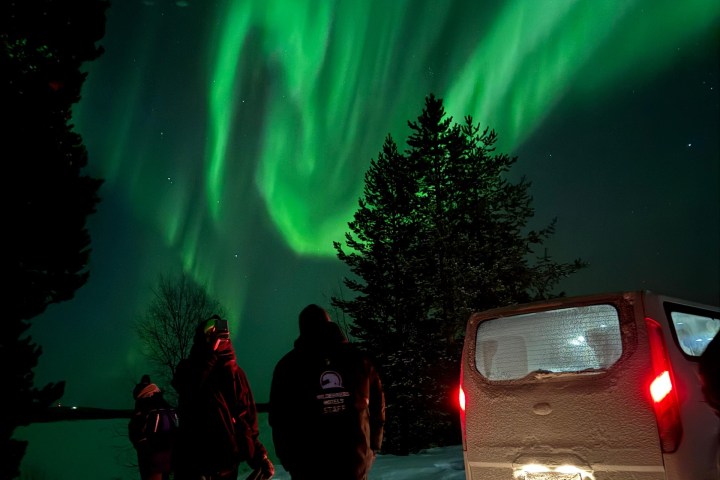 Three people watch green northern lights next to a snowy van in a forested area.