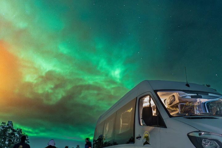 People near a white van under vibrant green Northern Lights and starry sky.