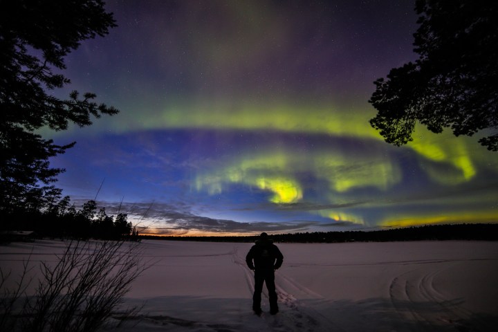 Person stands on snow gazing at vibrant aurora borealis under a starry sky.