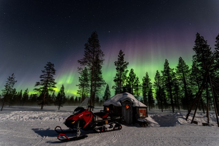 Aurora Borealis over snowy forest with a snowmobile and cabin in foreground.