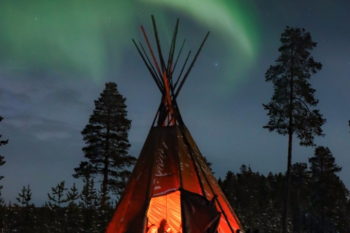 A tipi glows warmly under the northern lights in a snowy forest at night.