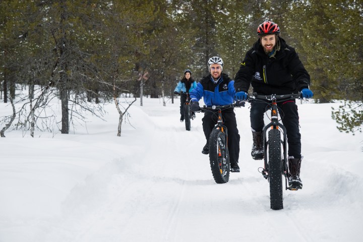 Three people riding fat bikes on a snowy forest trail, wearing winter gear and helmets.