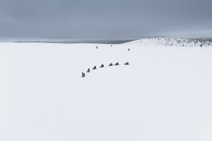 Snowmobiles in a line crossing vast snowy landscape under cloudy sky.