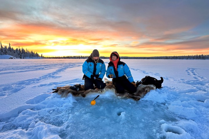 Two people ice fishing on a frozen lake at sunset with forest in background.