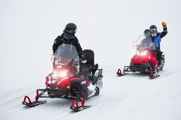 Two snowmobiles on a snowy terrain, riders wearing helmets, one waving.