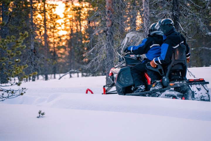 Two people ride a snowmobile through a snowy forest at sunset.