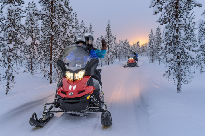 Two people on snowmobiles riding through snow-covered forest at sunset.