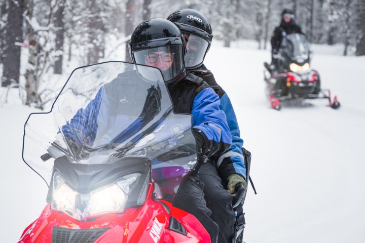 Two people riding a snowmobile in a snowy forest, with another rider in the background.