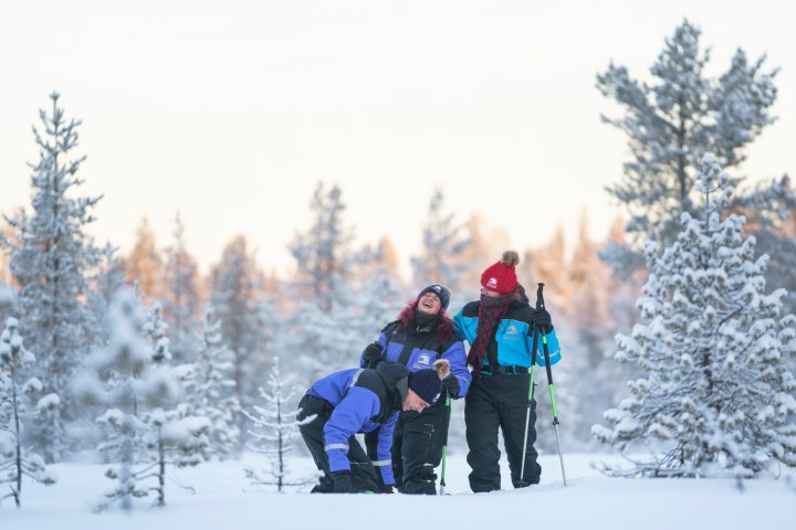 Three people in winter clothing laugh in a snowy forest, with snow-covered trees around.