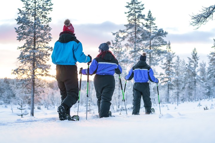 Three people in winter clothing snowshoeing through snowy forest at sunrise.
