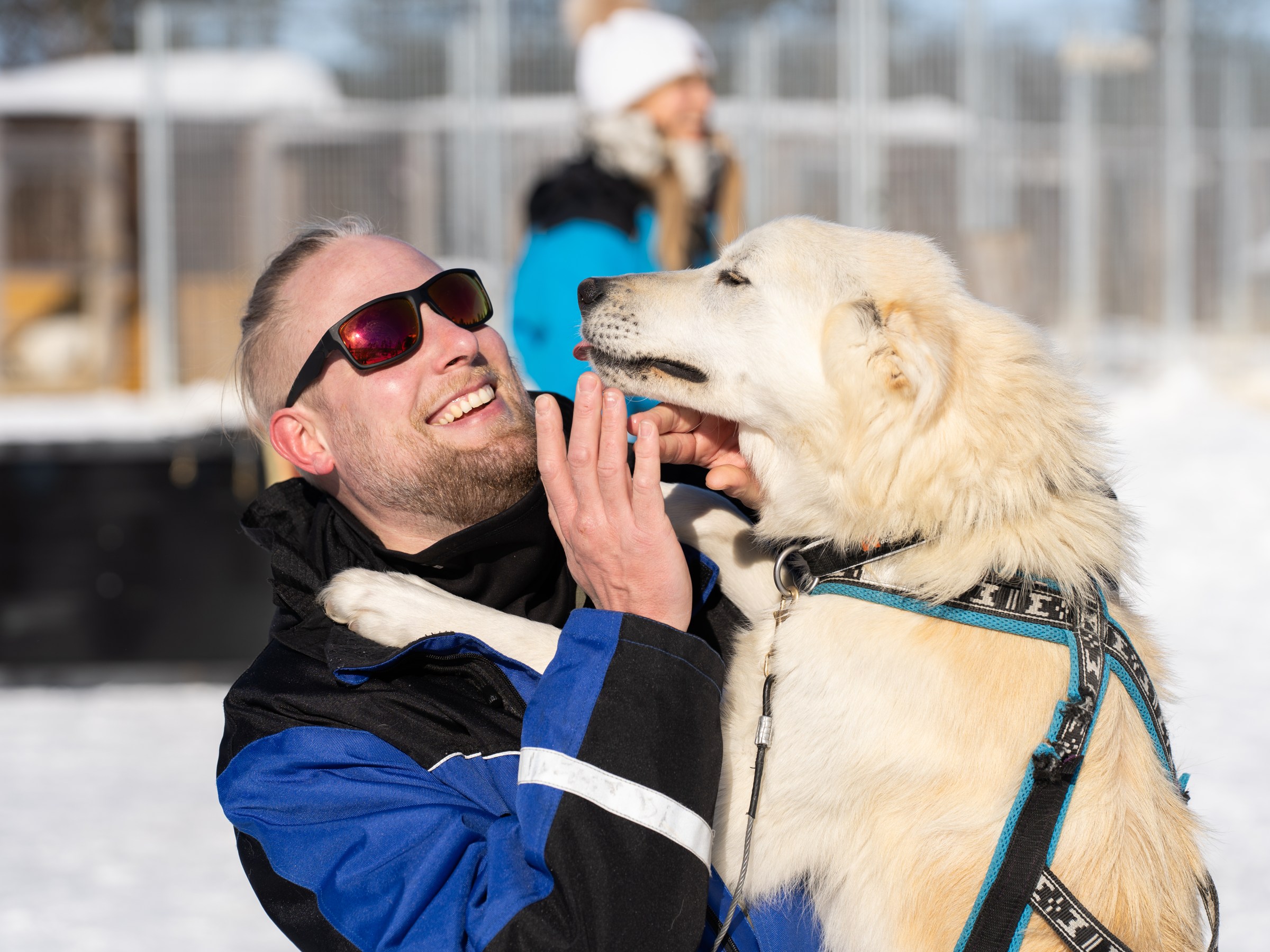 Man in sunglasses playing with a white dog in snowy outdoor setting.
