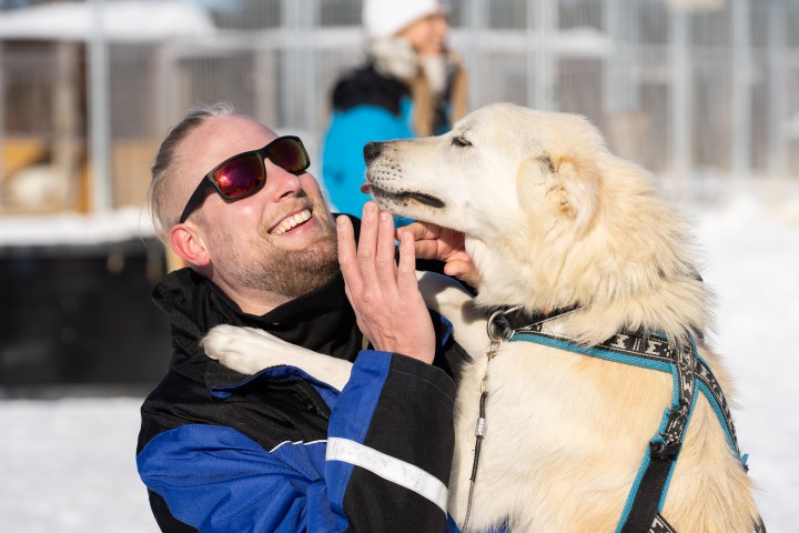 Man in sunglasses playing with a white dog in snowy outdoor setting.