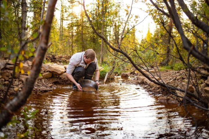 Person crouched by stream, panning for gold in a forested area with autumn foliage.