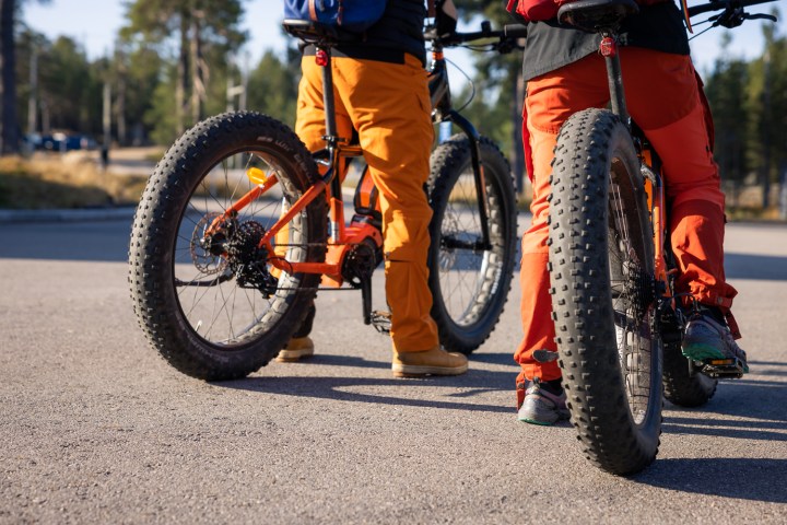Two cyclists in orange outfits on fat tire bikes stopped on a paved road.