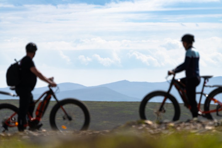 Two cyclists with mountain bikes on a hilltop, cloudy sky and distant mountains in the background.