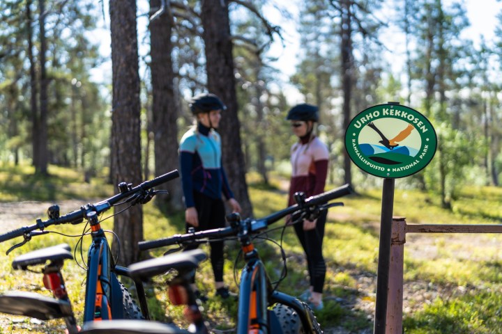 Two cyclists and bikes near Urho Kekkonen National Park sign in a forest.