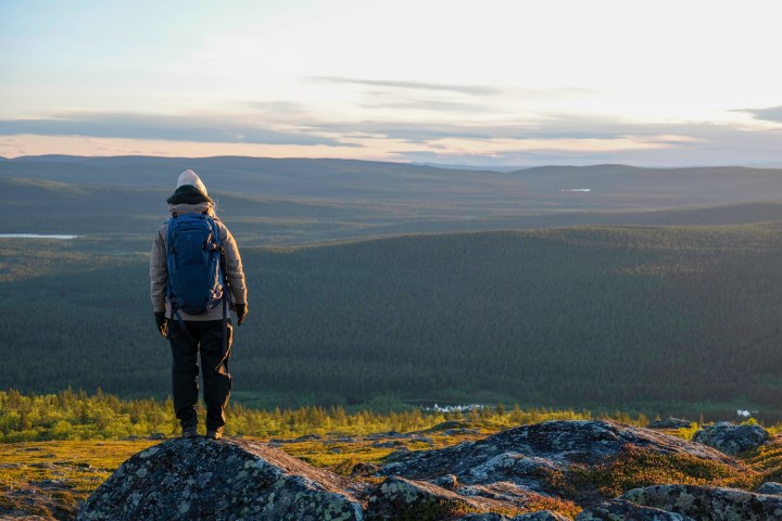 Person with backpack stands on rocky hill, overlooking forested landscape at sunset.