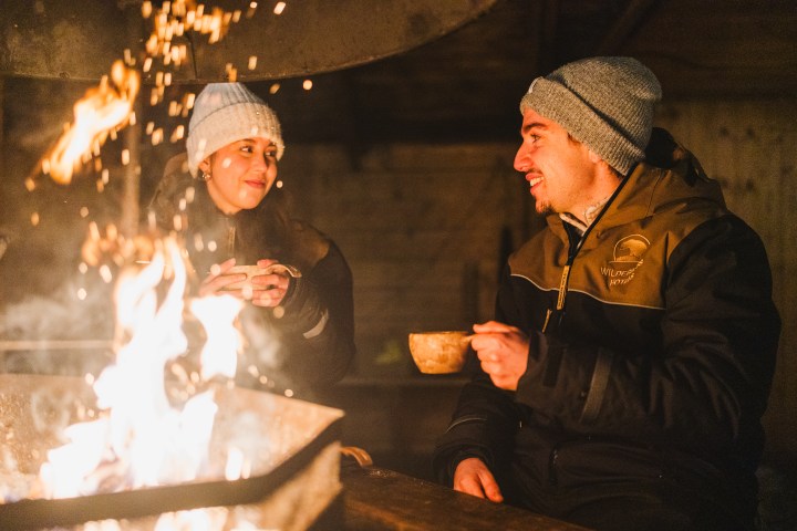 Two people in winter clothes enjoying drinks by a warm fire.