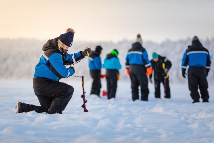 Person kneeling with ice auger, others standing on snowy field.
